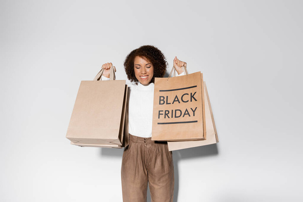 delighted african american woman with closed eyes and curly hair holding shopping bags with black friday lettering on grey - Fotografie, Obrázek