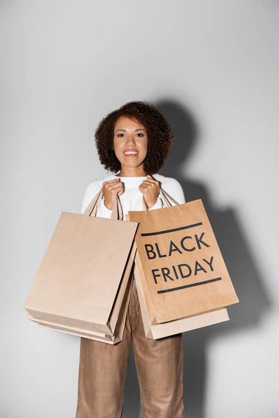 positive young african american woman holding shopping bags with black friday lettering on grey - 写真・画像