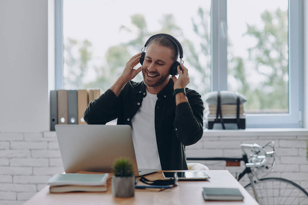 Happy young man in headphones using laptop while sitting at his working place in office - Фото, изображение
