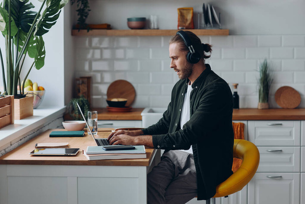 Confident man in headphones working on laptop while sitting at the kitchen counter at home - Foto, immagini