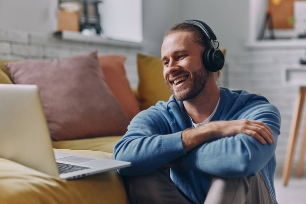 Handsome man in headphones looking at laptop and smiling while sitting on the floor at home - Foto, imagen