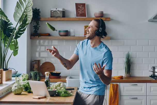 Happy young man in headphones tossing up a green pepper while preparing food at the kitchen - Fotoğraf, Görsel