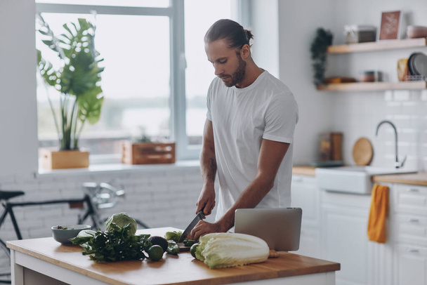 Confident young man cutting veggies while standing at the kitchen island at home - Foto, Bild