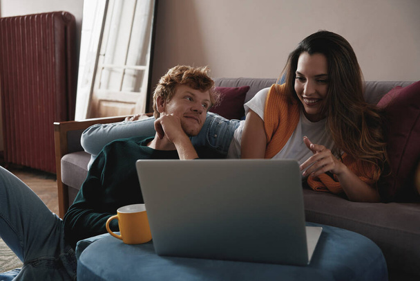 Beautiful young couple using laptop and smiling while relaxing at home together - Foto, imagen