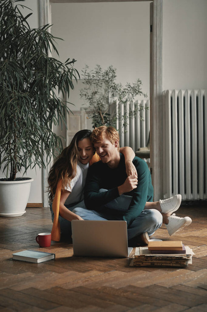 Young loving couple using laptop and smiling while sitting on the floor at home together - Foto, Imagen
