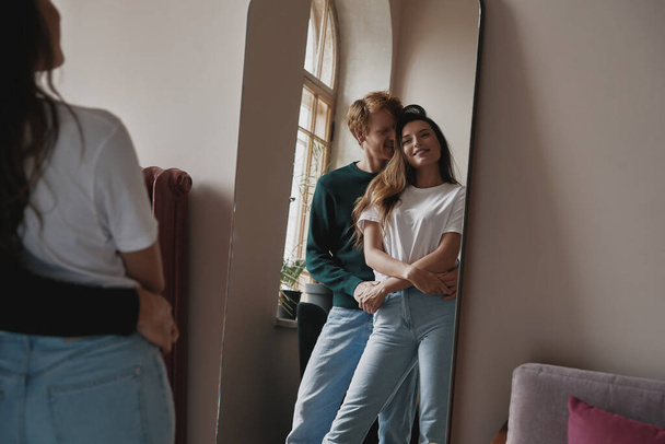 Happy young couple embracing while looking at their reflection in the mirror at home - Фото, зображення