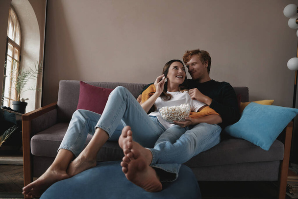 Happy young couple enjoying popcorn while relaxing on the couch at home - 写真・画像