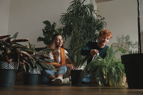 Beau jeune couple arrosant des plantes d'intérieur tout en étant assis sur le sol à la maison - Photo, image