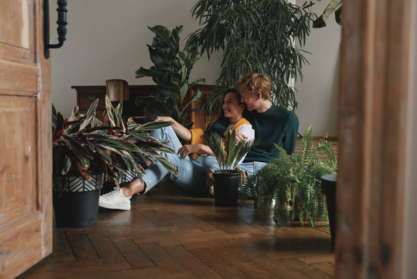 Beautiful young couple watering houseplants while sitting on the floor at home - Фото, изображение