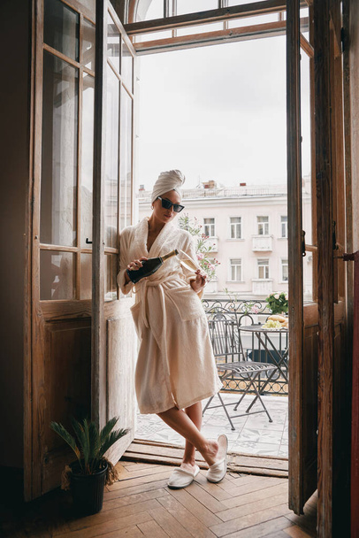 Full length of beautiful woman in bathrobe pouring champagne while leaning on the balcony door - Foto, Imagen