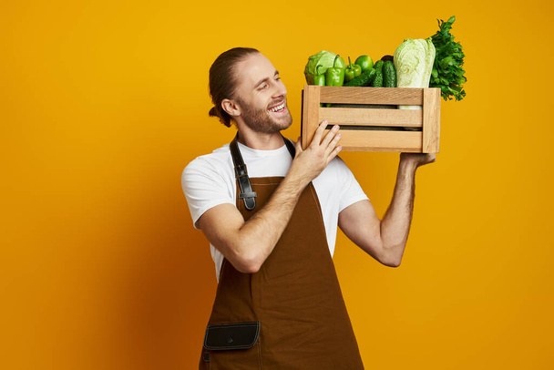 Cheerful young man in apron carrying wooden crate with fresh veggies against yellow background - Foto, Bild