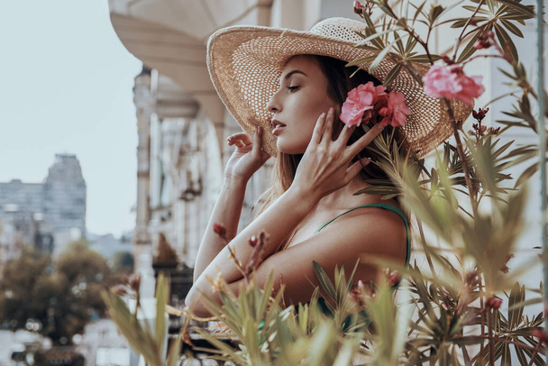 Attractive young woman in elegant hat holding flowers near face while standing on the balcony - Photo, image