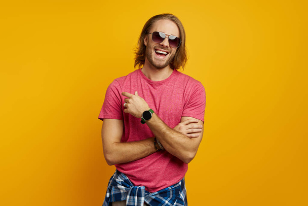 Handsome young man pointing away and smiling while standing against yellow background - Фото, изображение