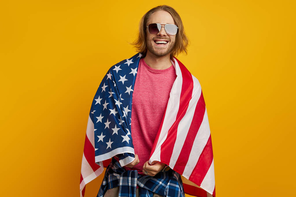 Bonito jovem coberto com bandeira americana olhando para a câmera e sorrindo contra fundo amarelo - Foto, Imagem