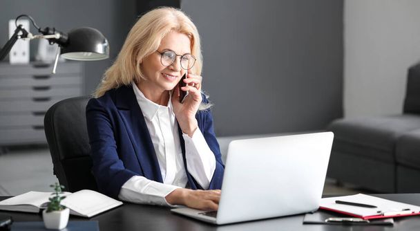 Mature businesswoman talking by phone while working on laptop in office - Foto, Bild