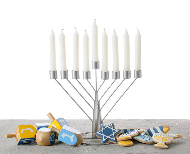 Menora mit Kerzen, Plätzchen und Dreidels zur Chanukka-Feier auf grauem Tisch vor weißem Hintergrund - Foto, Bild