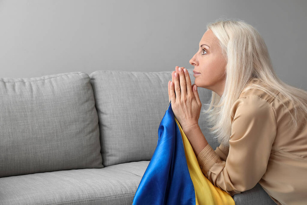 Mujer madura con bandera de Ucrania rezando cerca de sofá en casa - Foto, Imagen