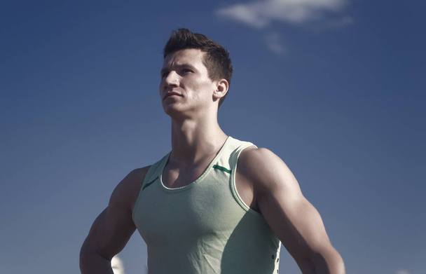 Athlete in green tshirt on sunny day. Sport and healthy lifestyle concept. Fitness model with muscular hands, biceps, triceps, strong muscles. Workout and training. Man bodybuilder posing on blue sky. - Фото, изображение