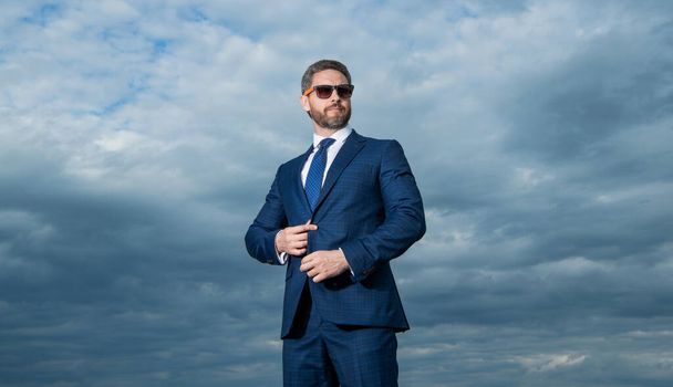 Manager wearing sunglasses sky background. Grizzled manager buttoning suit jacket. Serious manager outdoors. - 写真・画像