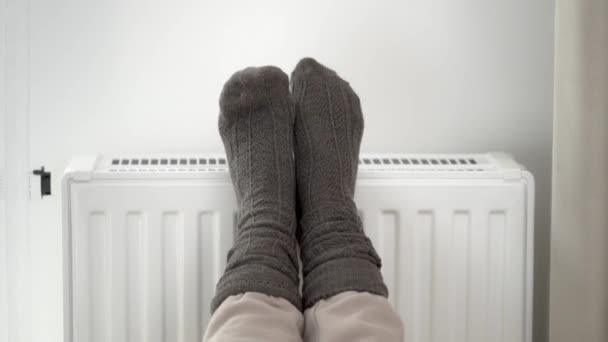 La femme a mis ses pieds gelés dans des chaussettes chaudes en laine sur le radiateur chaud du chauffage de la maison. Limiter la consommation d'énergie pendant la période froide de chauffage hivernal pendant la crise énergétique mondiale. - Séquence, vidéo