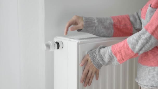 Una mujer toca el radiador frío de un calentador de casa con sus manos congeladas y aumenta la potencia de calefacción por el valor máximo. Limitar el consumo de energía durante la temporada de calefacción. La crisis energética. - Imágenes, Vídeo