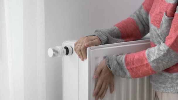 Uma mulher toca o radiador frio de um aquecedor doméstico com as mãos congeladas e aumenta o poder de aquecimento pelo valor máximo. Limitar o consumo de energia durante a estação de aquecimento. A crise energética. - Filmagem, Vídeo