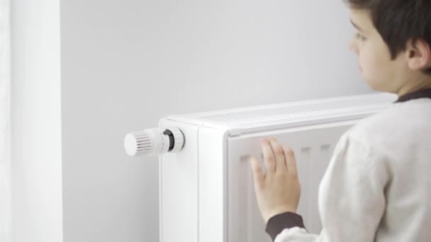 A criança toca o radiador frio do aquecedor doméstico com as mãos congeladas e aumenta seu poder de aquecimento pelo valor máximo para aquecer. O rapaz abraça o aquecedor. Aquecimento durante a crise energética. - Filmagem, Vídeo