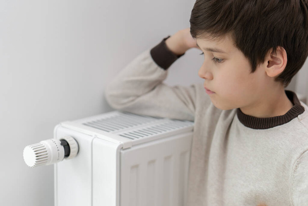 The child hugs the radiator of the home heater with his hands to keep warm. Heating of housing during the energy crisis in the winter cold season. Restrictions and savings of gas and electricity. - Fotografie, Obrázek