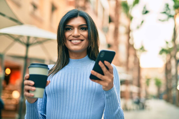 Jonge Spaanse vrouw die smartphone gebruikt en een kop koffie drinkt in de stad - Foto, afbeelding