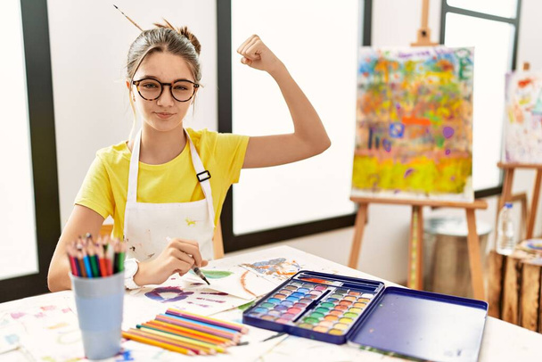 jong brunette tiener in kunst studio sterk persoon tonen arm spier, zelfverzekerd en trots op de macht  - Foto, afbeelding