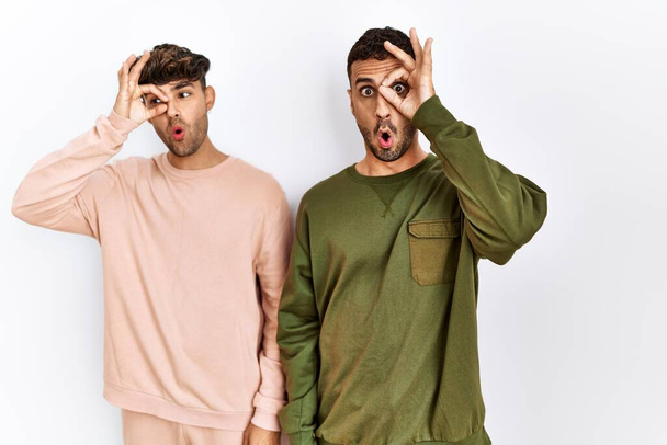 Young gay couple standing over isolated white background doing ok gesture shocked with surprised face, eye looking through fingers. unbelieving expression.  - Photo, Image