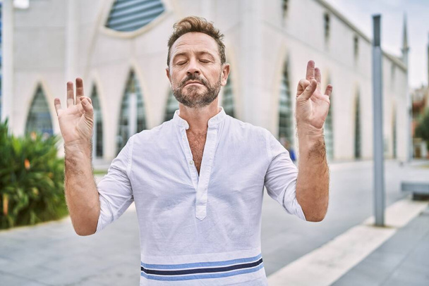 Şehirdeki orta yaşlı adam rahatlar ve gözleri kapalı gülümser parmaklarıyla meditasyon jesti yapar. yoga konsepti.  - Fotoğraf, Görsel