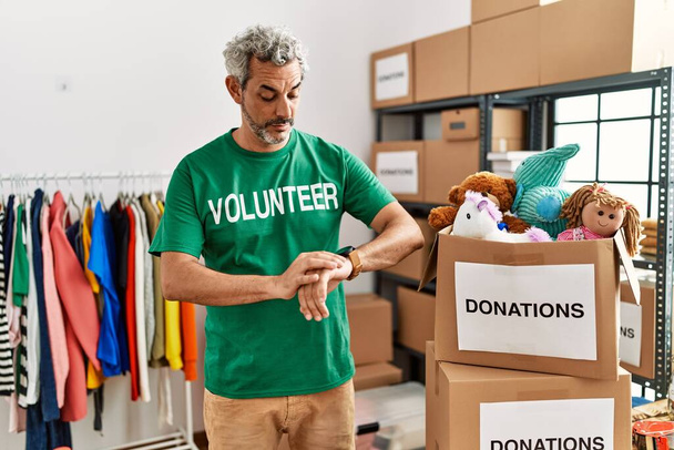 Middle age hispanic man wearing volunteer t shirt at donations stand checking the time on wrist watch, relaxed and confident  - Photo, Image
