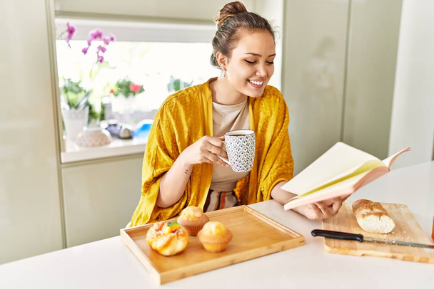 Jonge mooie Spaanse vrouw ontbijten leesboek in de keuken - Foto, afbeelding