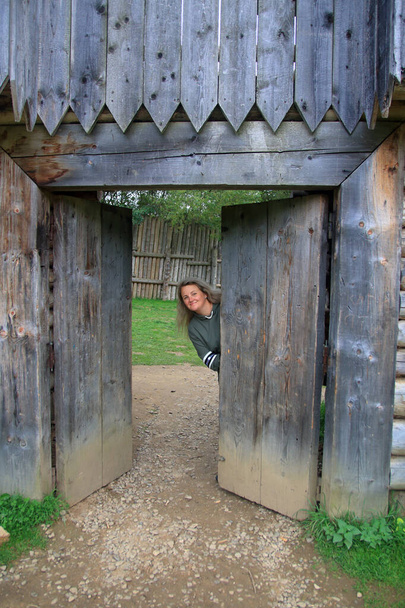 Η φωτογραφία τραβήχτηκε στα Καρπάθια Όρη κοντά στην είσοδο ενός αντιγράφου ενός ελεύθερα επισκέψιμου φρουρίου. Η φωτογραφία δείχνει ένα κορίτσι να κρυφοκοιτάζει πίσω από μια παλιά ξύλινη πύλη.. - Φωτογραφία, εικόνα