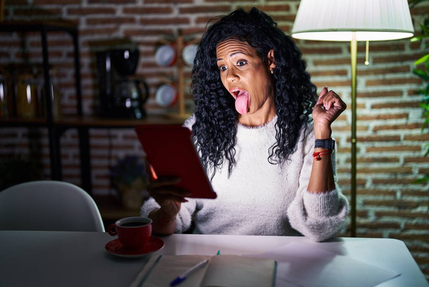 Middle age hispanic woman using touchpad sitting on the table at night sticking tongue out happy with funny expression. emotion concept.  - Фото, изображение