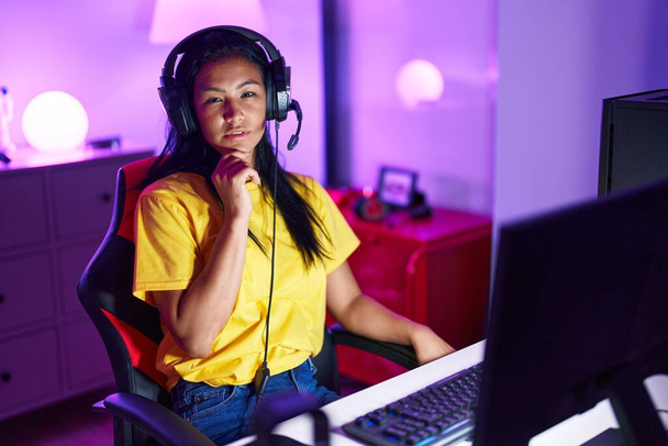 Young beautiful latin woman streamer sitting on table with serious expression at gaming room - Photo, image