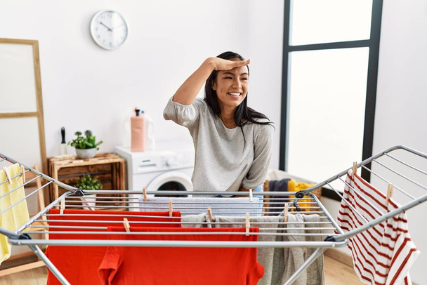 Young hispanic woman putting fresh laundry on clothesline very happy and smiling looking far away with hand over head. searching concept.  - Photo, Image