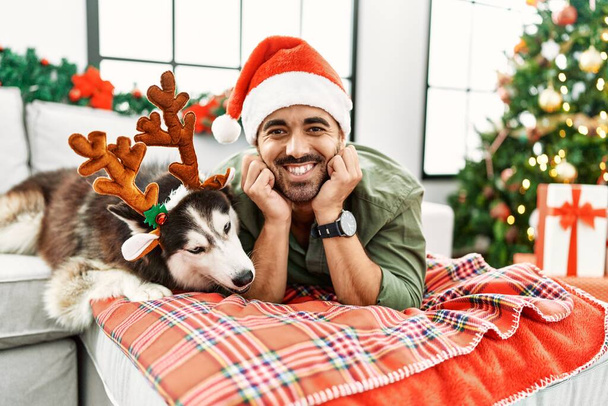 Junger hispanischer Mann liegt mit Hund auf Sofa und trägt Rentierhut am Weihnachtsbaum - Foto, Bild