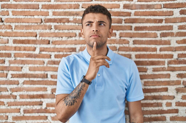 Brasilianischer junger Mann steht über Ziegelmauer und denkt konzentriert über Zweifel nach, mit dem Finger am Kinn und schaut fragend nach oben  - Foto, Bild