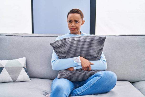 Femme afro-américaine assise sur le canapé à la maison étreignant l'oreiller sceptique et nerveux, fronçant les sourcils contrariés en raison de problèmes. personne négative.  - Photo, image