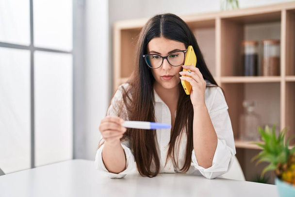 妊娠検査の結果を持っている若いブルネットの女性は、顔に深刻な表情でリラックスした電話で話しています。カメラを見ているだけで.  - 写真・画像