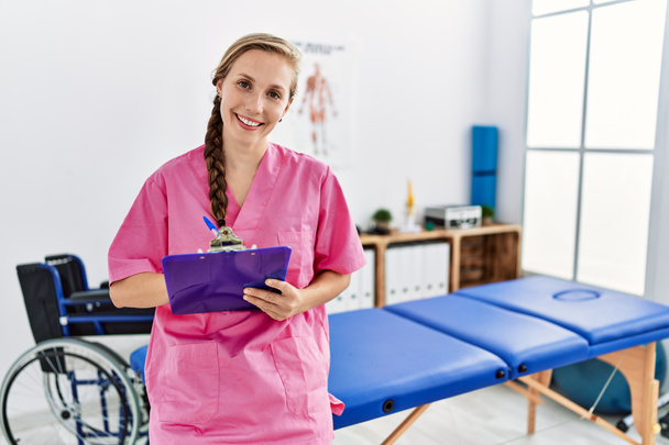 Jonge blanke vrouw draagt fysiotherapeut uniform schrijven op checklist staan bij fysiotherapie kliniek - Foto, afbeelding