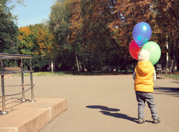 Üç parlak balonu olan küçük bir çocuk parktaki ağaçlara bakıyor. Arka plan - Fotoğraf, Görsel