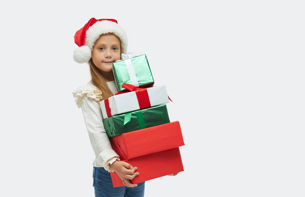 Χριστουγεννιάτικο παιδί. Χαριτωμένο κοριτσάκι στο καπέλο Σάντα Claus κρατώντας υψηλή στοίβα από πολύχρωμα κουτιά δώρων σε λευκό φόντο - Φωτογραφία, εικόνα