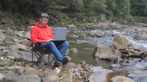 A man works on a laptop near a stream in nature. Concept of freelancing, digital nomad or remote office. 4K - Felvétel, videó