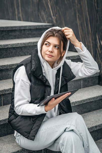 A young woman student against the background of steps stands with a digital tablet in her hands, the concept of technology and education. - Foto, Bild