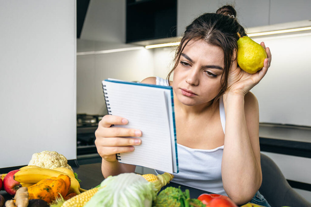 Egy fiatal nő a konyhában egy jegyzettömbbel a zöldségek között, a főzés, a diéta és az egészséges táplálkozás fogalma.. - Fotó, kép