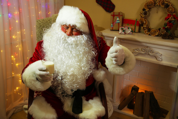 Санта-Клаус сидит с молочным стаканом в удобном кресле у камина дома
 - Фото, изображение