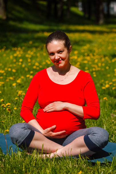 Exercice de yoga de grossesse femme enceinte faisant asana Sukhasana pose de yoga facile tenant son abdomen à l'extérieur sur la pelouse d'herbe avec des pissenlits en été - Photo, image